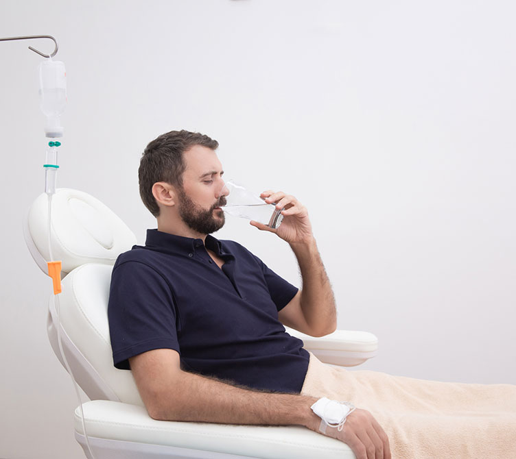 Sick, dehydrated or hangover patient man receiving vitamin IV infusion drip and drinking glass of water in hospital or beauty salon. Healthcare and medicine concept.